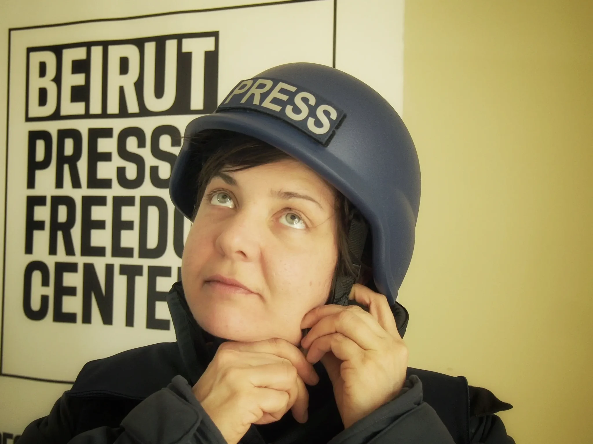 En journalist med blå hjälm märkt "PRESS" justerar hakbandet och blickar uppåt. I bakgrunden syns en skylt med texten "Beirut Press Freedom Center".