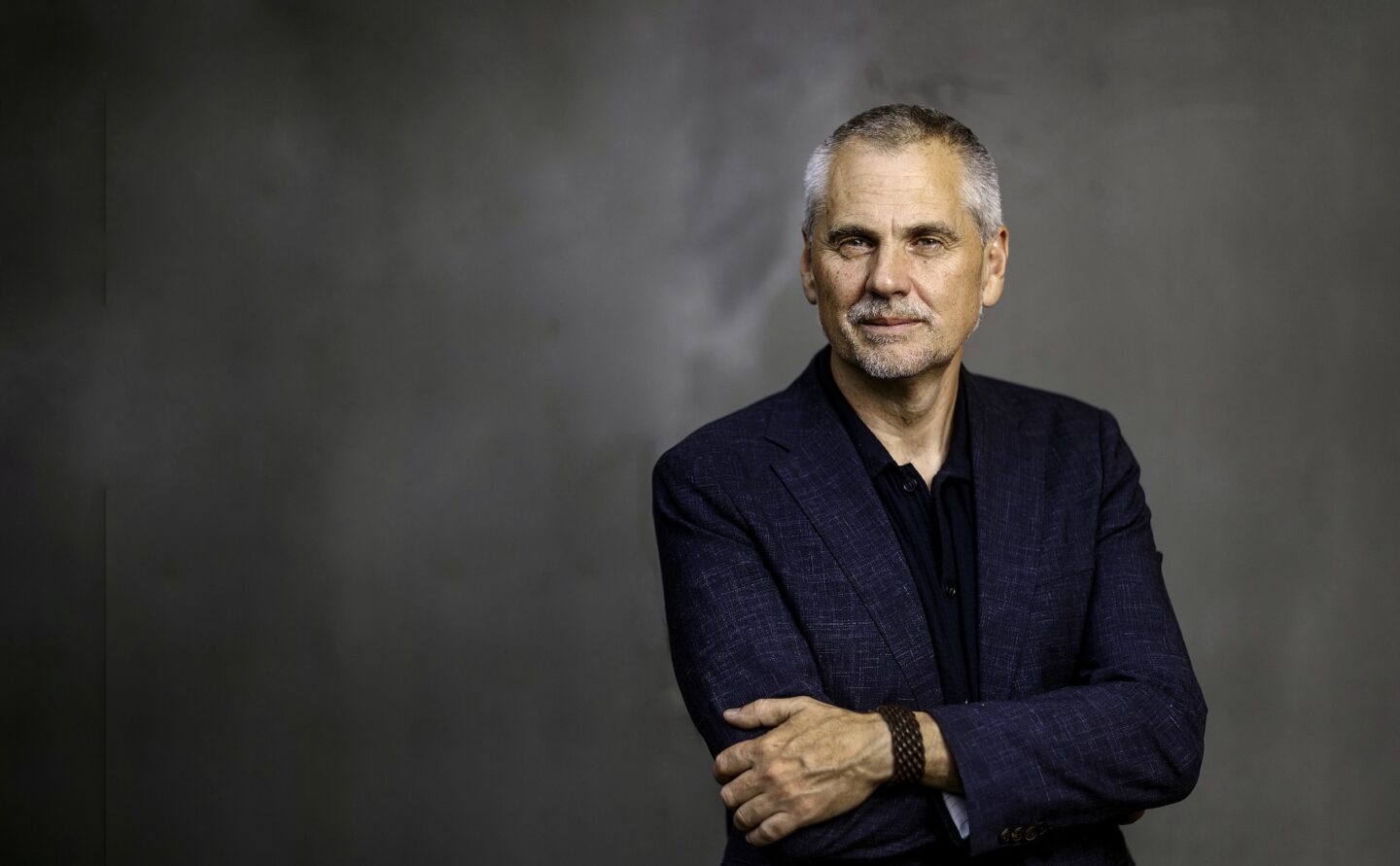 En medelålders man med grått hår och kort skägg står framför en neutral grå bakgrund. Han bär en mörkblå kavaj över en mörk skjorta och har armarna i kors. Uttrycket är självsäkert och lätt leende. Ljussättningen är mjuk och professionell, med fokus på ansiktet.
