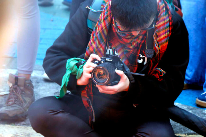 En person i närbild sitter på marken och håller i en svart Nikon DSLR-kamera. Personen bär en mörk jacka, en färgglad röd- och grönrutig sjal som täcker ansiktet och ett grönt tygband knutet runt handleden. I bakgrunden syns benen på förbipasserande personer på en gata.