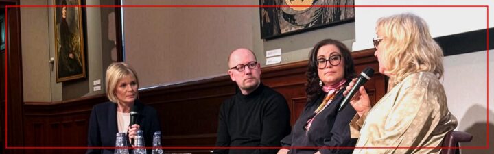 Ett fotografi av en paneldiskussion med fyra personer sittande på en scen. Från vänster till höger: En blond kvinna i mörkblå kostym håller en penna och ett block vid ett litet bord med vattenflaskor; en skallig man med glasögon bär en svart polotröja; en mörkhårig kvinna med glasögon, svart klädsel och rosa scarves håller en mikrofon; och längst till höger, en blond kvinna i guldskjorta talar in i en mikrofon med båda händerna. Bakgrunden visar mörk träpanel och oljemålningar. En röd ram omger bilden.