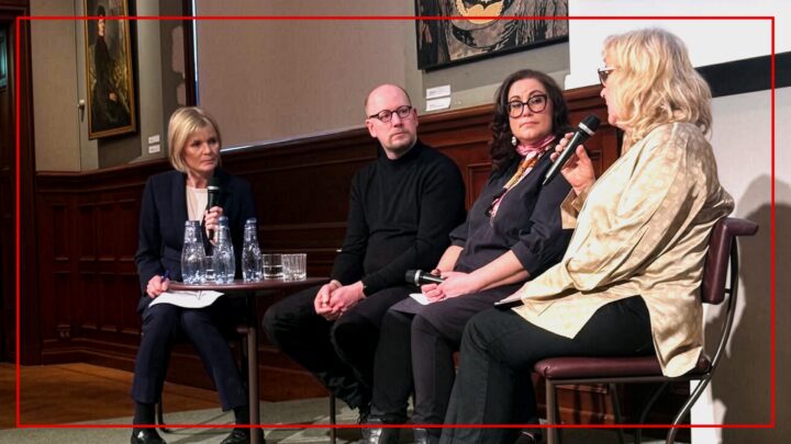 Ett fotografi av en paneldiskussion med fyra personer sittande på en scen. Från vänster till höger: En blond kvinna i mörkblå kostym håller en penna och ett block vid ett litet bord med vattenflaskor; en skallig man med glasögon bär en svart polotröja; en mörkhårig kvinna med glasögon, svart klädsel och rosa scarves håller en mikrofon; och längst till höger, en blond kvinna i guldskjorta talar in i en mikrofon med båda händerna. Bakgrunden visar mörk träpanel och oljemålningar. En röd ram omger bilden.