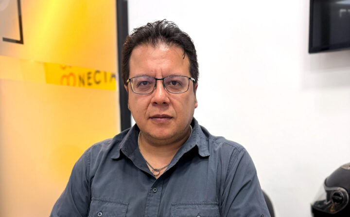 En mansperson som tittar rakt fram, iklädd en grå skjorta och glasögon. Han har mörkt hår och en ring-formad halsband på sig. I bakgrunden finns en gul och vit yta.