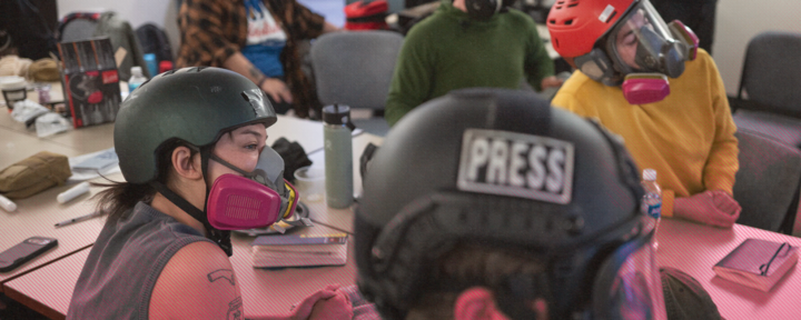 I förgrunden syns ryggarna på två journalister som sitter vid ett bord. Personen längst till höger bär en svart hjälm med en text som lyder: "PRESS". Båda personerna har på sig andningsmasker och skyddsglasögon. I bakgrunden syns ytterligare två personer som också bär skyddsutrustning. Bordet de sitter vid är fyllt med andra föremål, såsom anteckningsböcker, vattenflaskor och pennor.