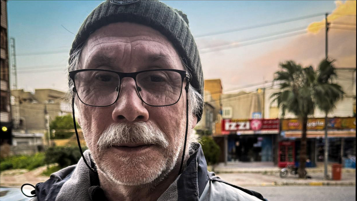 Närbild av en äldre man med glasögon och stickad mössa som tittar rakt in i kameran. I bakgrunden syns en gata i en stadsmiljö med låga byggnader, skyltar, en palm och en motorcykel; ljuset är dämpat, som vid skymning eller efter regn.