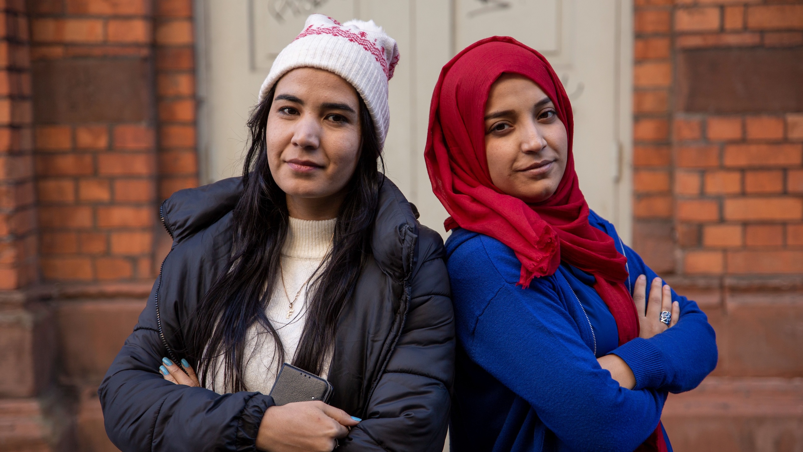 Två unga afghanska kvinnor med armarna i kors står framför en tegelbyggnad. En bär vintermössa och den andra röd hijab.