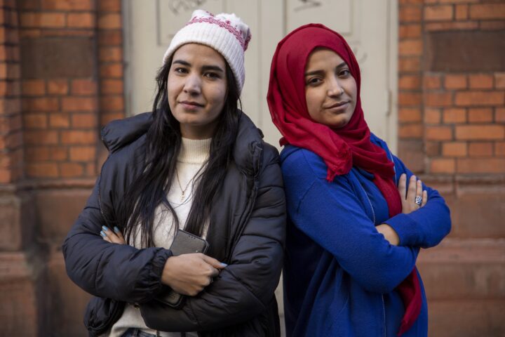 Två unga afghanska kvinnor med armarna i kors står framför en tegelbyggnad. En bär vintermössa och den andra röd hijab.